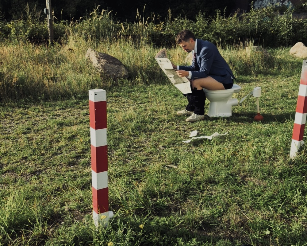 A suited actor reading a newspaper while sitting on a toilet outdoors